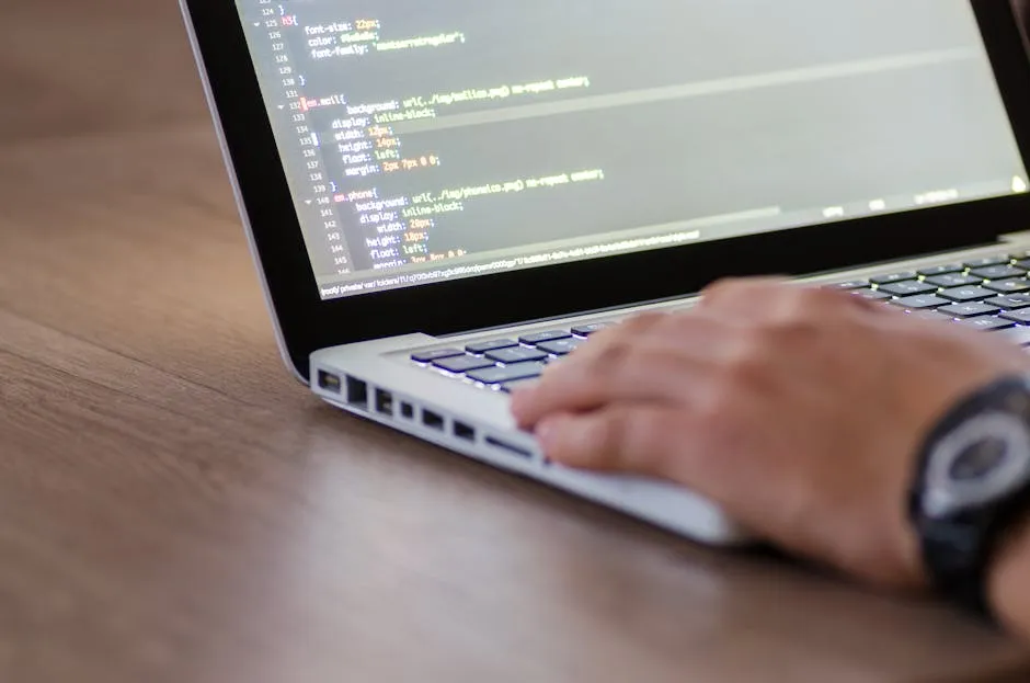 Close-up of a developer's hands at a keyboard with a monitor displaying code in the background, suggesting active investigation of a software defect